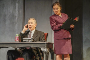 A male actor sits at a desk on the phone while a female actors stand next to him listening in on the conversation.