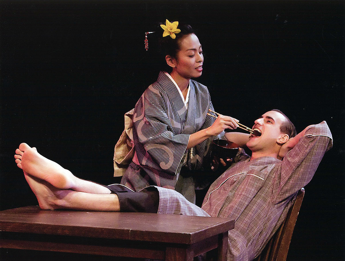 A actor in a traditional kimono feeds another sitting at a desk with feet popped up. She is feeding the other with chopsticks.