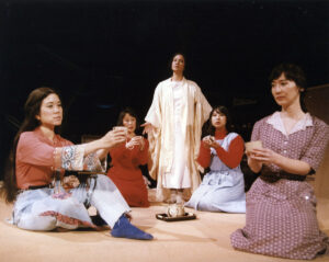 Five woman actors hold tea cups, sitting in a circle and look off into the distance.